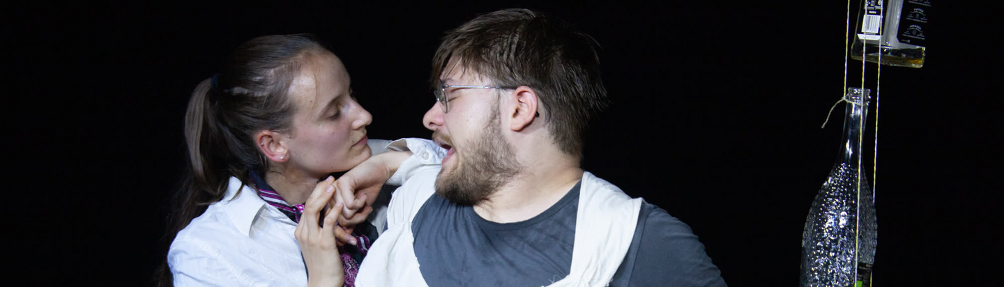 Das Bild zeigt zwei junge Männer vor einem blauen Hintergrund mit hellem Lichteffekt, der an Theaterbeleuchtung erinnert. Beide tragen Brillen und schauen überrascht und gespannt direkt in die Kamera. Der Mann links hat schulterlanges braunes Haar, einen Bart und trägt ein rot-weiß gemustertes T-Shirt. Der Mann rechts hat die Haare nach hinten gebunden, trägt einen Vollbart und ein weißes T-Shirt mit roten Streifen. Er zieht seine Brille leicht nach unten, um über den Rand hinwegzusehen, als könne er nicht glauben, was er sieht. Ihre Gesichtsausdrücke sind überrascht, begeistert und wirken, als wären sie mitten in einer spannenden Theaterszene.